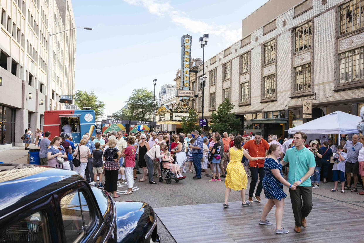 Unos bailarines realizan una presentación junto a autos de la década de 1940 afuera del Centro de Artes Escénicas Coronado antes de una proyección del primer episodio de la nueva serie de Amazon Prime "A League of Their Own", en Rockford, Illinois, el 2 de julio de 2022. (Taylor Glascock/The New York Times)
