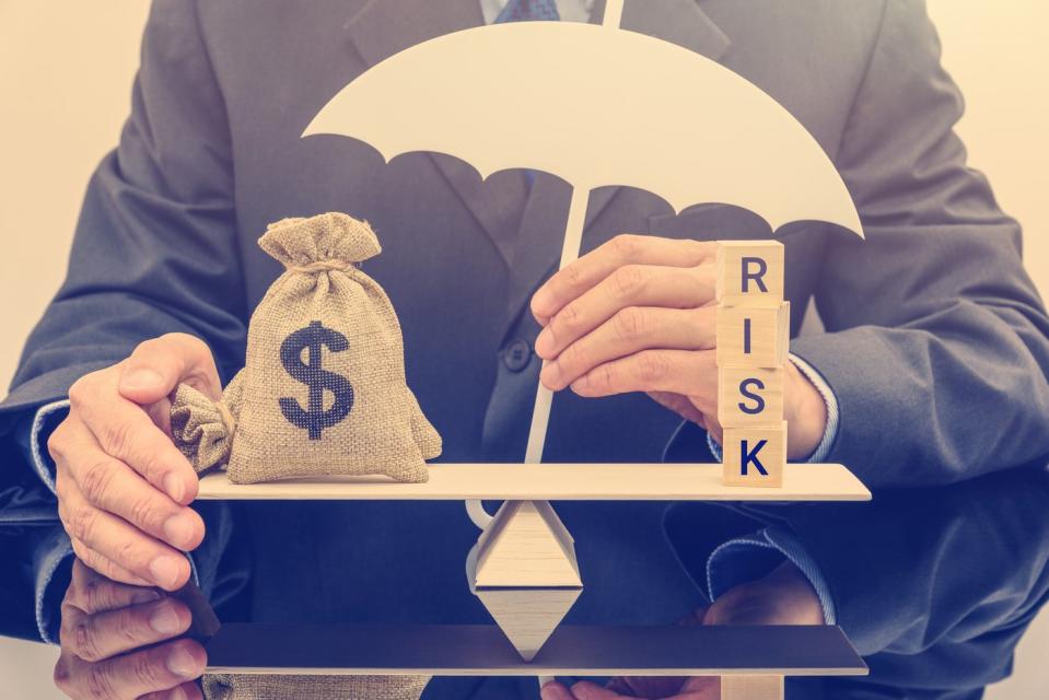 A man with the word risk and a bag of money balanced in front of him in a simple balance with an umbrella over the whole.