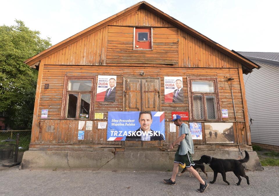 A man walks his dogs outside a home with campaign posters showing two candidates in Poland's weekend presidential election, Polish President Andrzej Duda and his challenger, Warsaw Mayor Rafal Trzaskowski, in Tykocin, Poland, on Tuesday July 7, 2020. Two bitter rivals are heading into a razor's-edge presidential runoff election Sunday in Poland that is seen as an important test of populism in Europe after a campaign that exacerbated a conservative-liberal divide in the country. (AP Photo/Czarek Sokolowski)