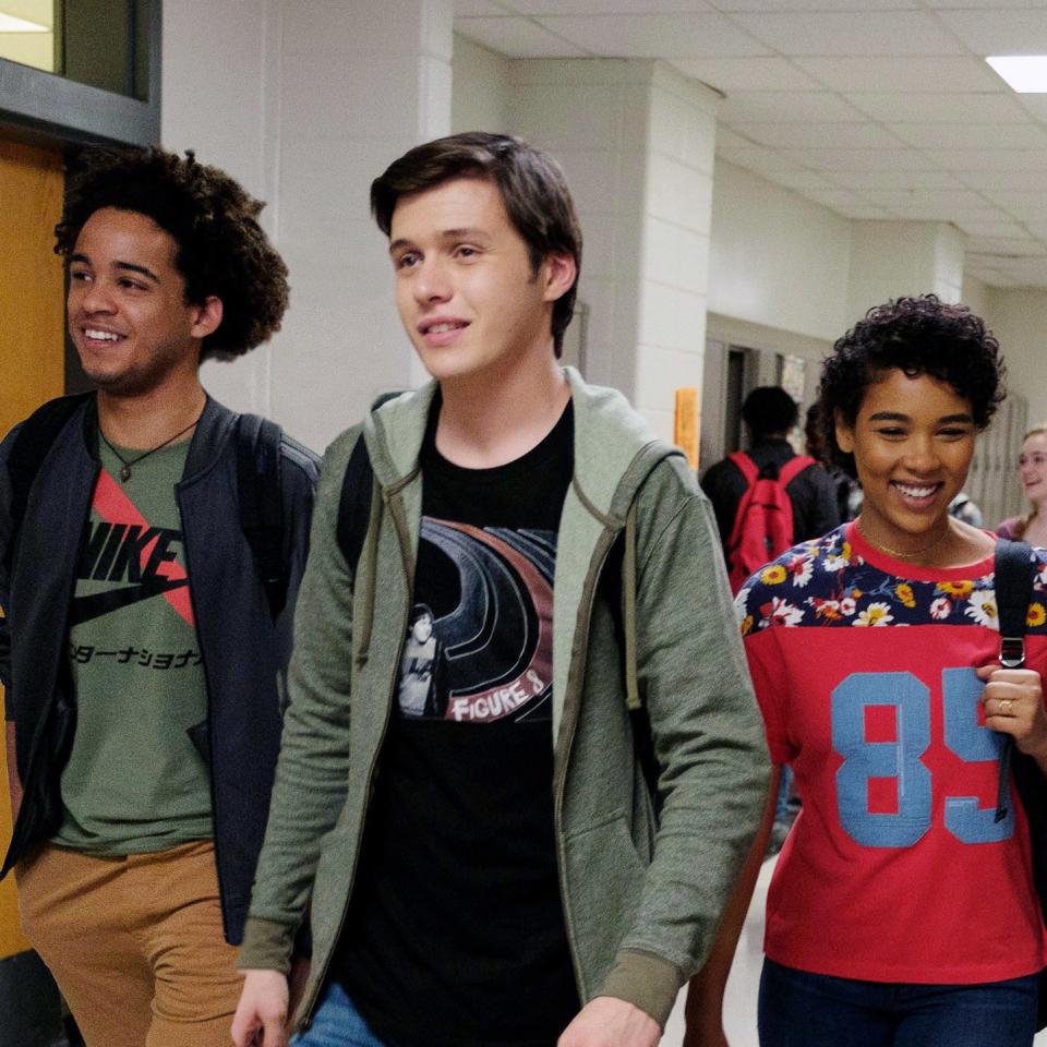 A group of four teens, Simon, Leah, Abby, and Nick, walk down a school hallway chatting and smiling. Simon wears a jacket, Leah holds a tote bag, Abby wears a jersey, and Nick wears a hoodie