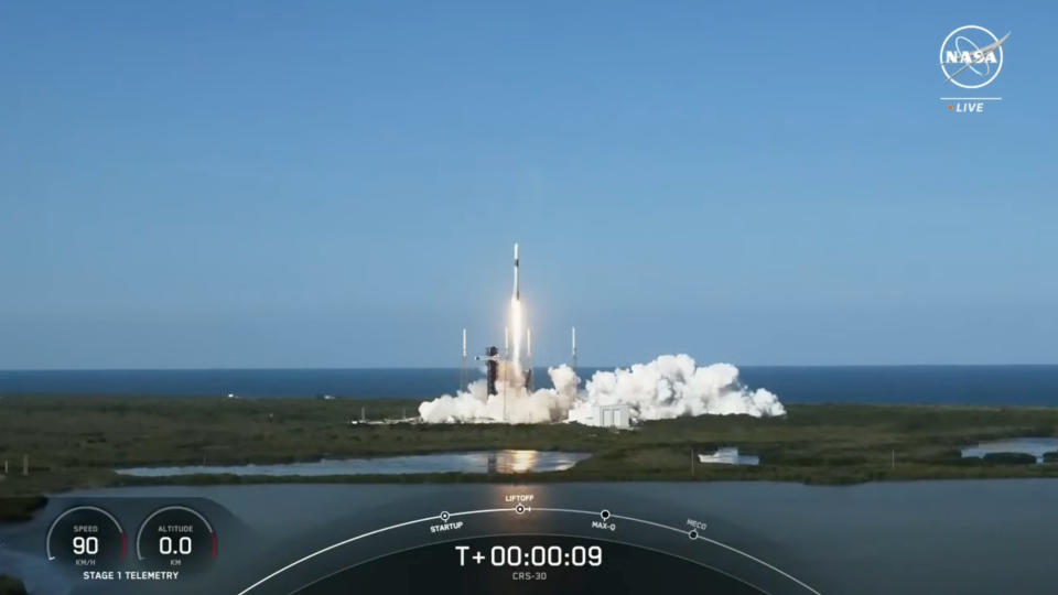 a black and white spacex rocket launches into a blue sky, with the ocean in the background