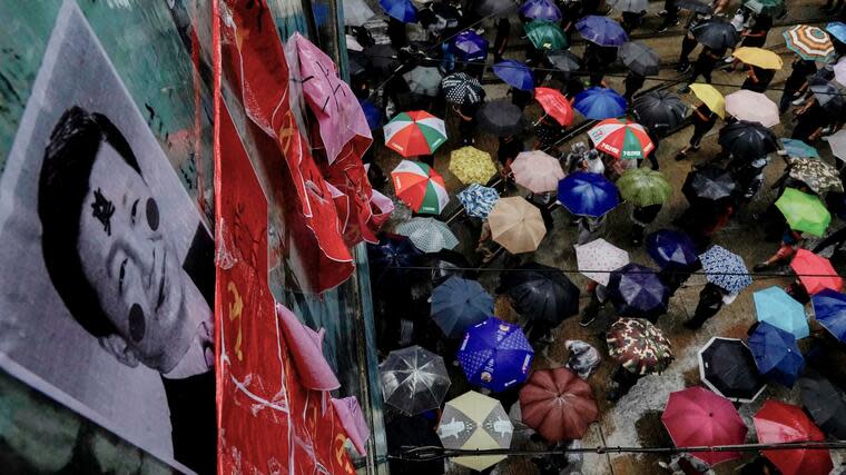 Hongkong: Demonstranten mit Regenschirmen marschieren an einer Fußgängerbrücke vorbei, worauf ein zerstörtes Bild des chinesischen Präsidenten Xi Jinping und kommunistische Parteifahnen hängen. Foto: dpa