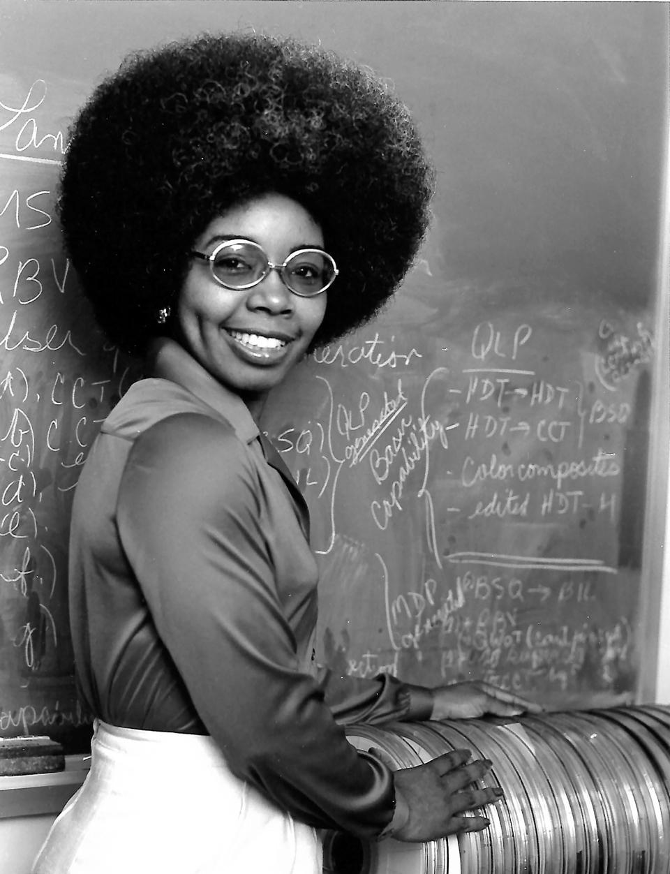 In this black and white photo, Valerie Thomas wears glasses and light pants while standing in front of a chalkboard.