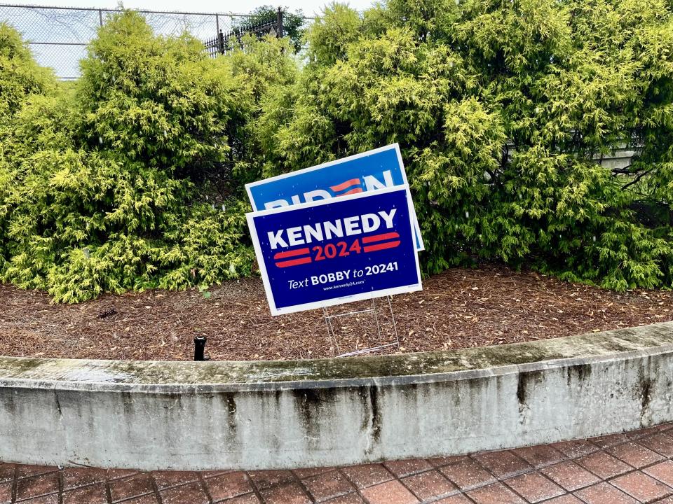 An RFK Jr. campaign sign is seen in front of a Biden-Harris one in a mulch bed along a brick sidewalk.