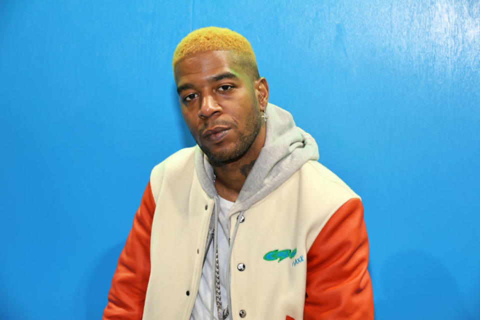Kid Cudi in varsity jacket sitting against a wall at an event