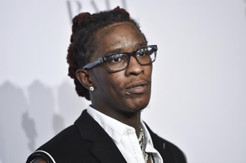 Young Thug posing with a blank expression, wearing thick-framed glasses and a black vest and white button-up shirt