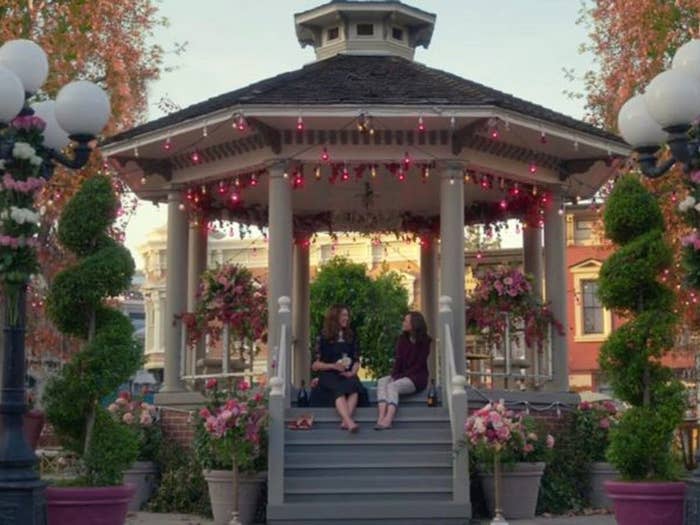 Lorelai and Rory from Gilmore Girls sit on the top step under a gazebo that&#39;s decorated with lights, town buildings are behind them