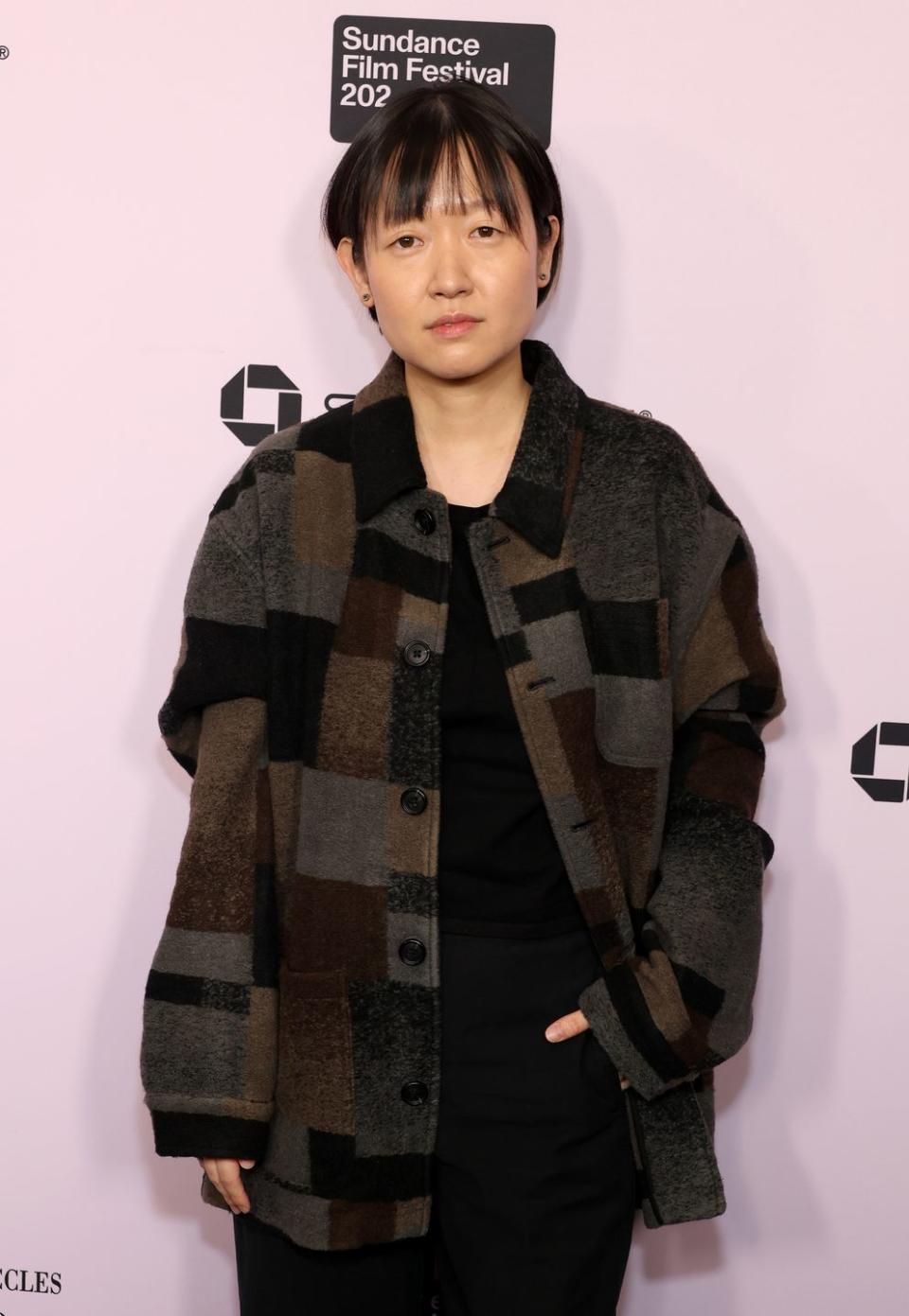 celine song looks at the camera, she wears a brown, black, and gray jacket over an all black outfit, behind her is a pink background with black logos