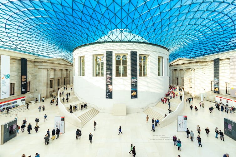 Inside the Great Court of the British Museum. A large, round, white central structure is covered in a glass roof, with people standing around in an expansive forecourt.