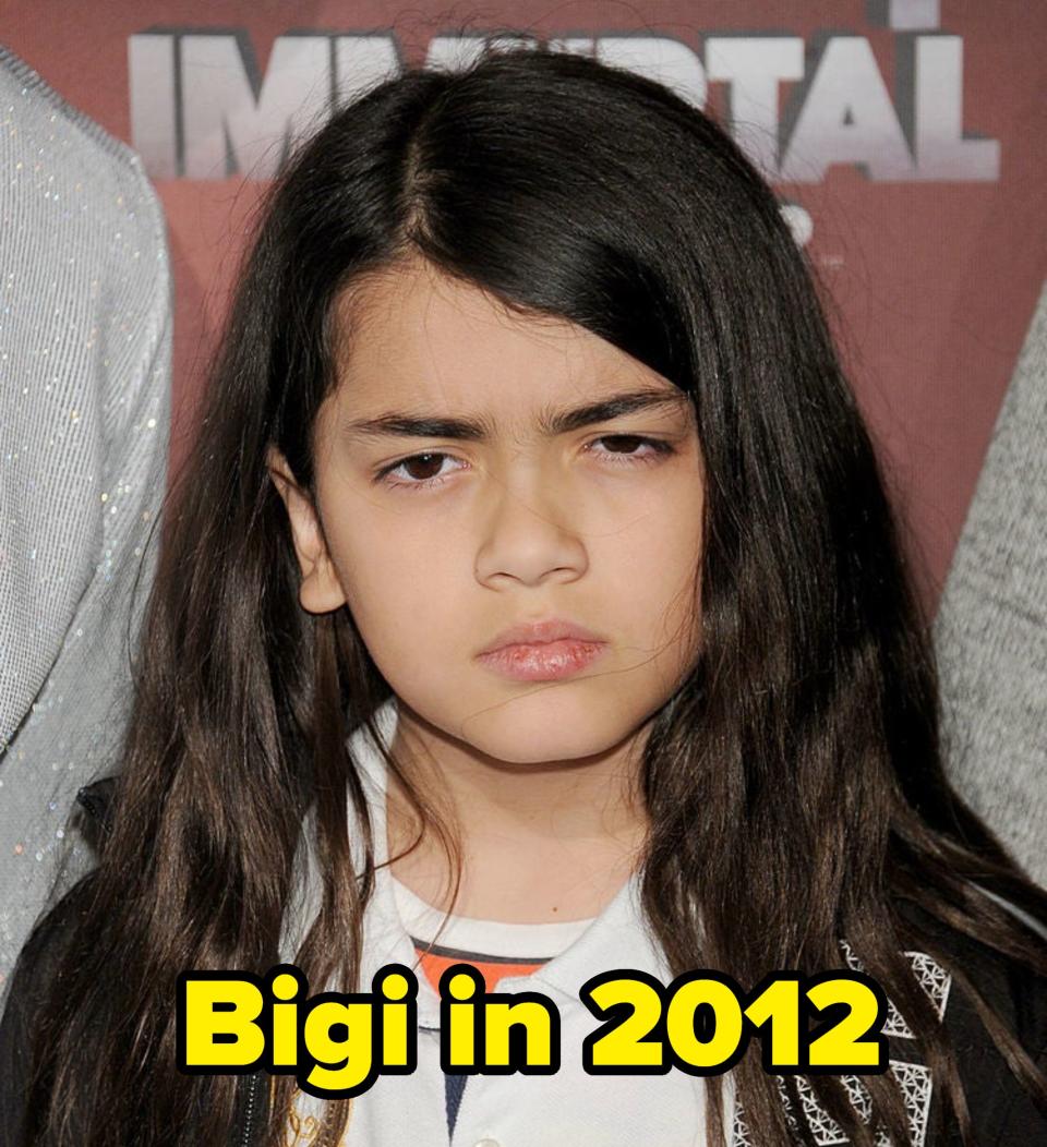 Child with a serious expression wearing a white shirt with badges, at a red carpet event