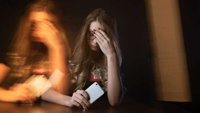 A girl holds her face in one hand and a cell phone in the other and is mirrored across the image