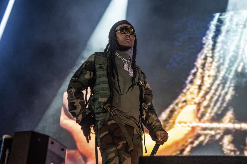 A man with long black hair wearing camouflage and holding a microphone on a stage