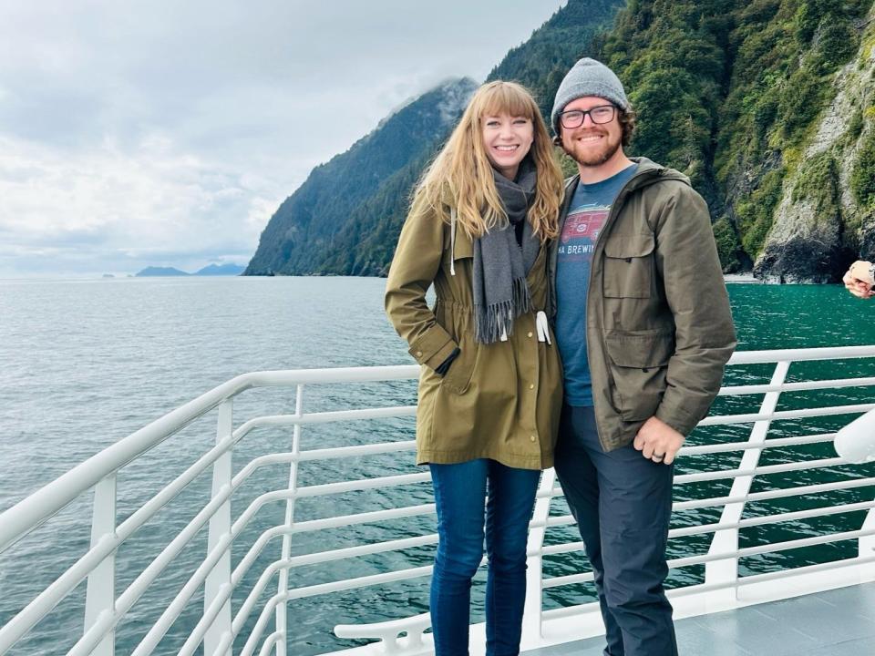 Emily and her husband smile on the deck of a boat, with greenery-covered mountains in the background.