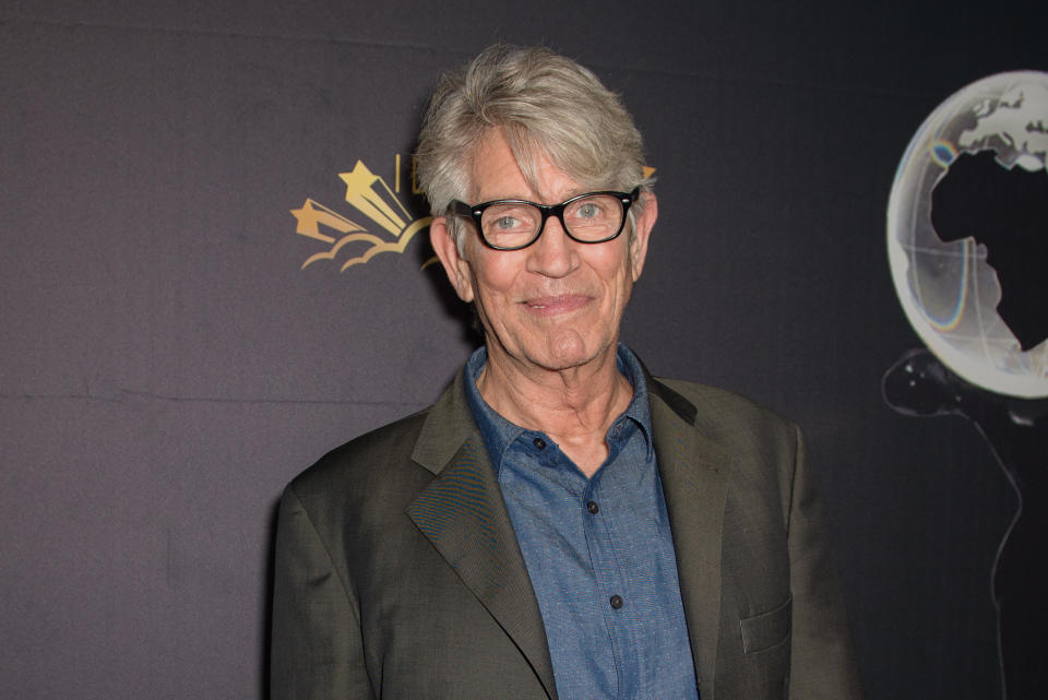 Eric Roberts is seen smiling at a red carpet event, wearing a casual blue shirt under a brown blazer and black-framed glasses