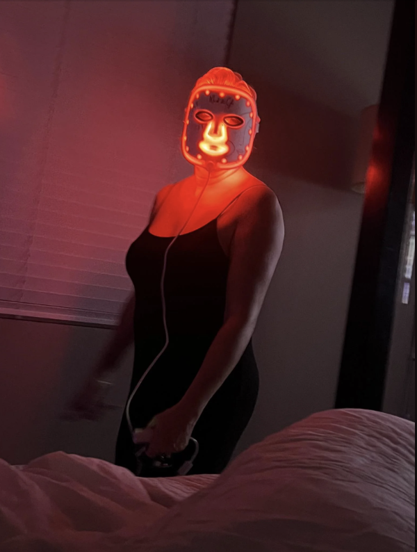Person in a fitted dress wearing a glowing face mask, standing near a bed in a dimly lit room