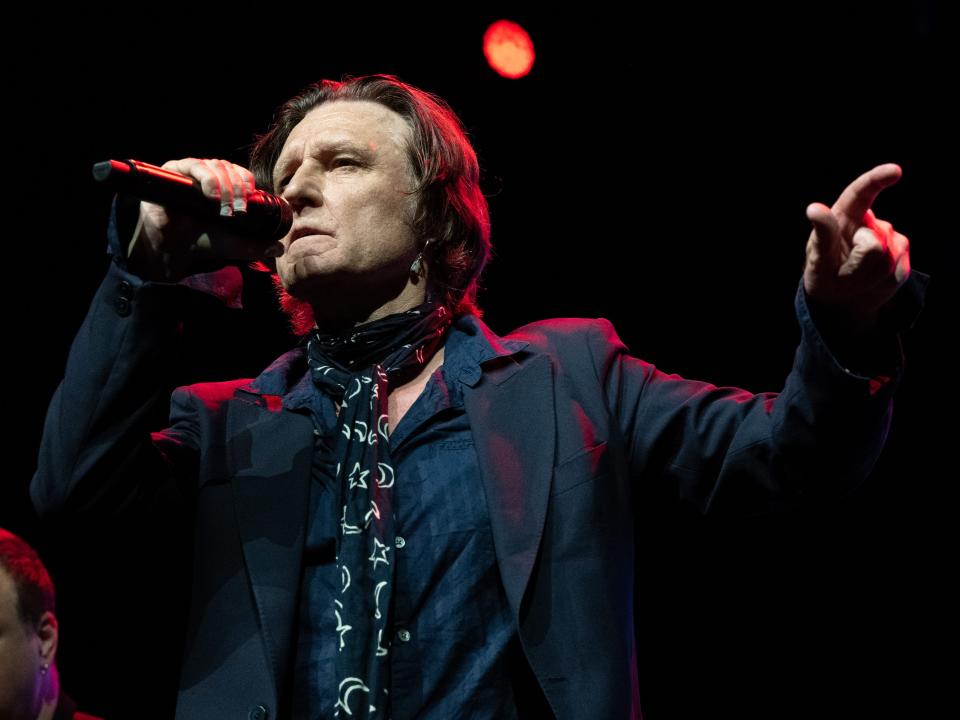 singer john waite onstage, holding up a microphone and gesturing with his left hand. he's wearing a navy shirt, black blazer, and black and white patterned scarf