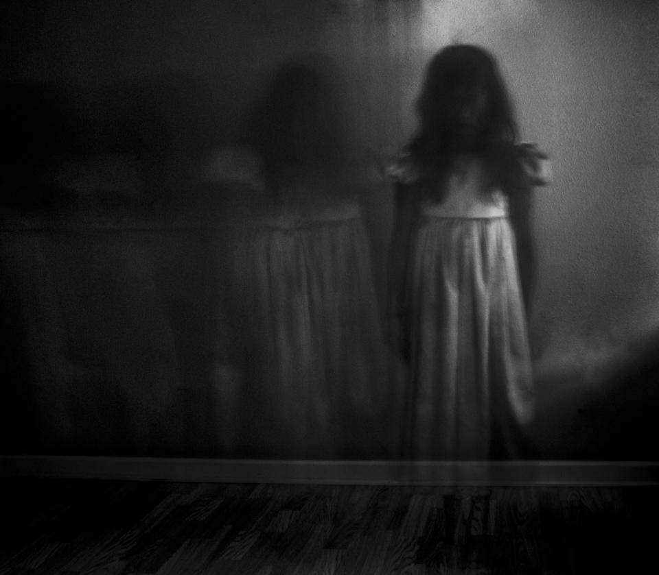 A girl in a white dress stands in a dimly lit room with her reflection on the wall. The atmosphere is dark and eerie