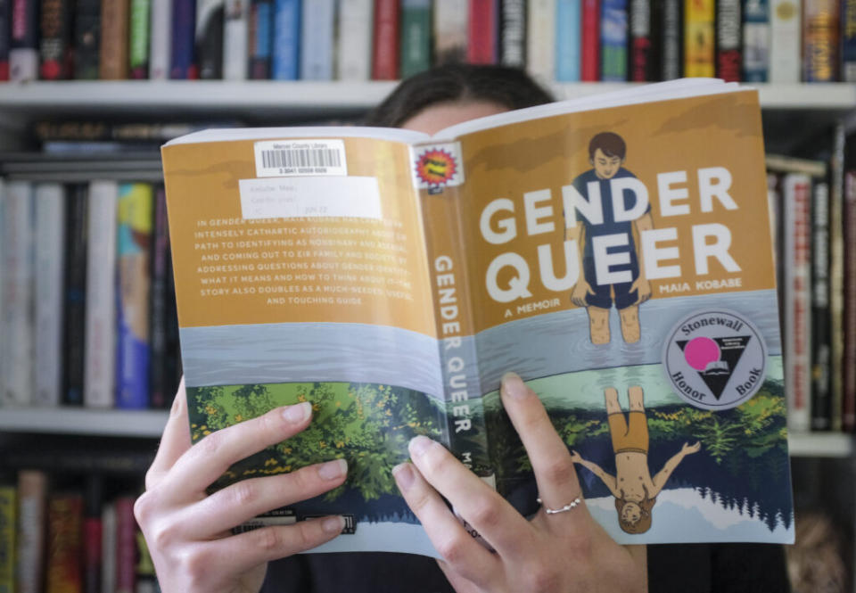 A person seated in a library reads the book "Gender Queer," with the book obscuring their face.
