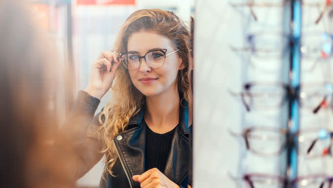 Smiling young woman trying on glasses on mirror in optician.