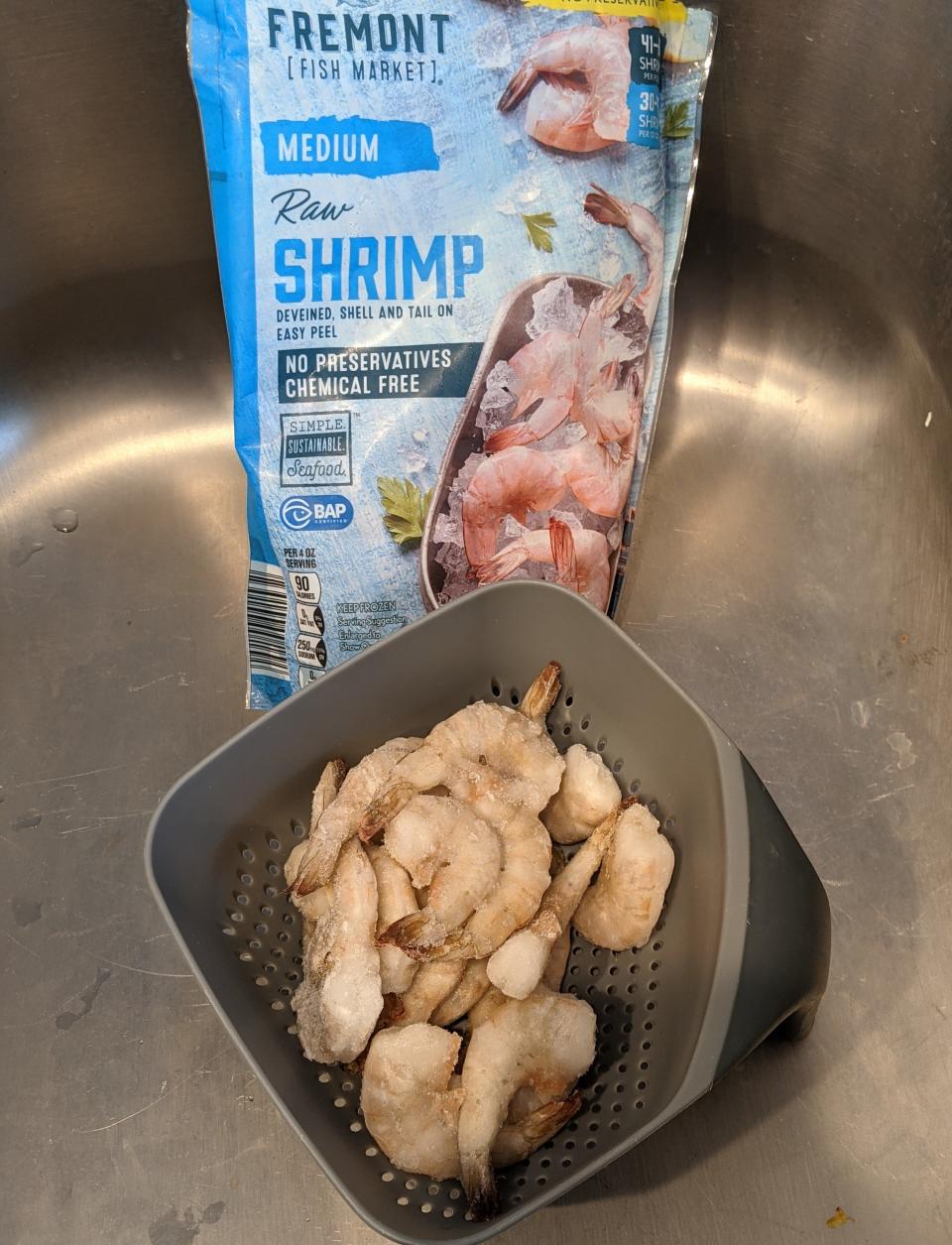 A colander full of defrosting shrimp in a sink with the blue original packaging from Aldi