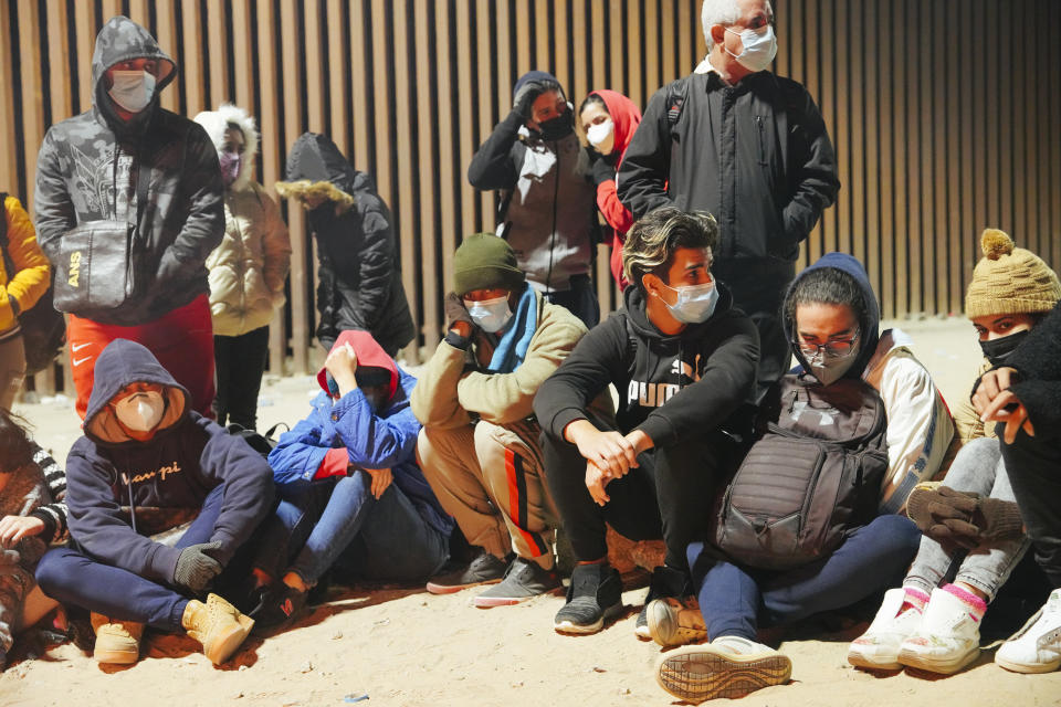 Asylum seekers from Cuba wearing masks and warm jackets sit on the dirt, with others standing.