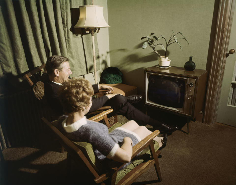 A man and woman sit in armchairs watching a vintage TV in a 1960s-style living room with a potted plant and a lamp