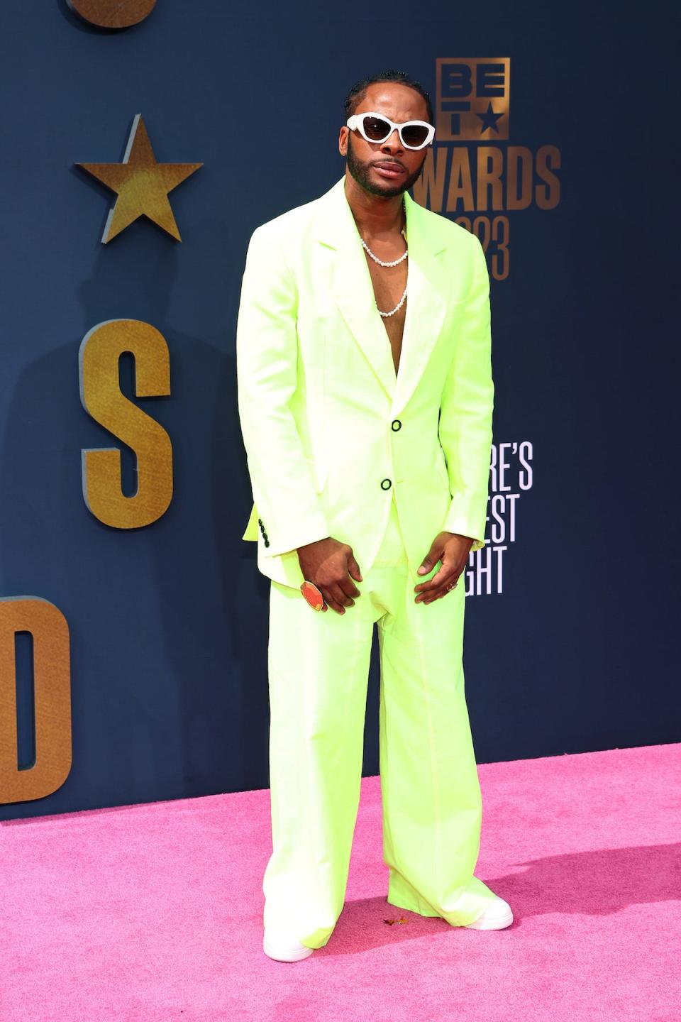 Dexta Daps poses on the red carpet at the 2023 BET Awards wearing a shirtless, neon green suit with two necklaces, white sneakers and sunglasses.