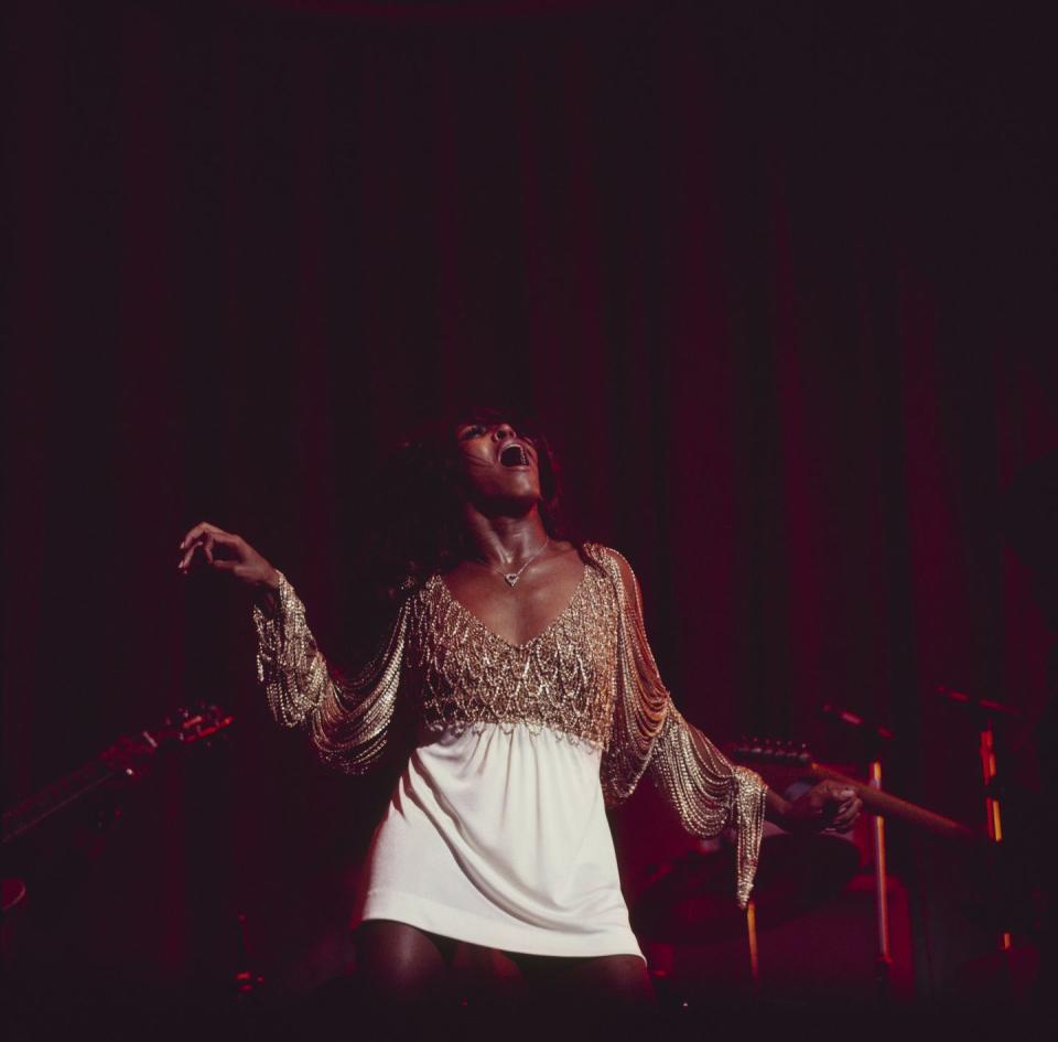 tina turner performs on stage, her head is lifted upward and her mouth open while singing, she is wearing a white skirt with a fringe top, her arms are extended at her sides