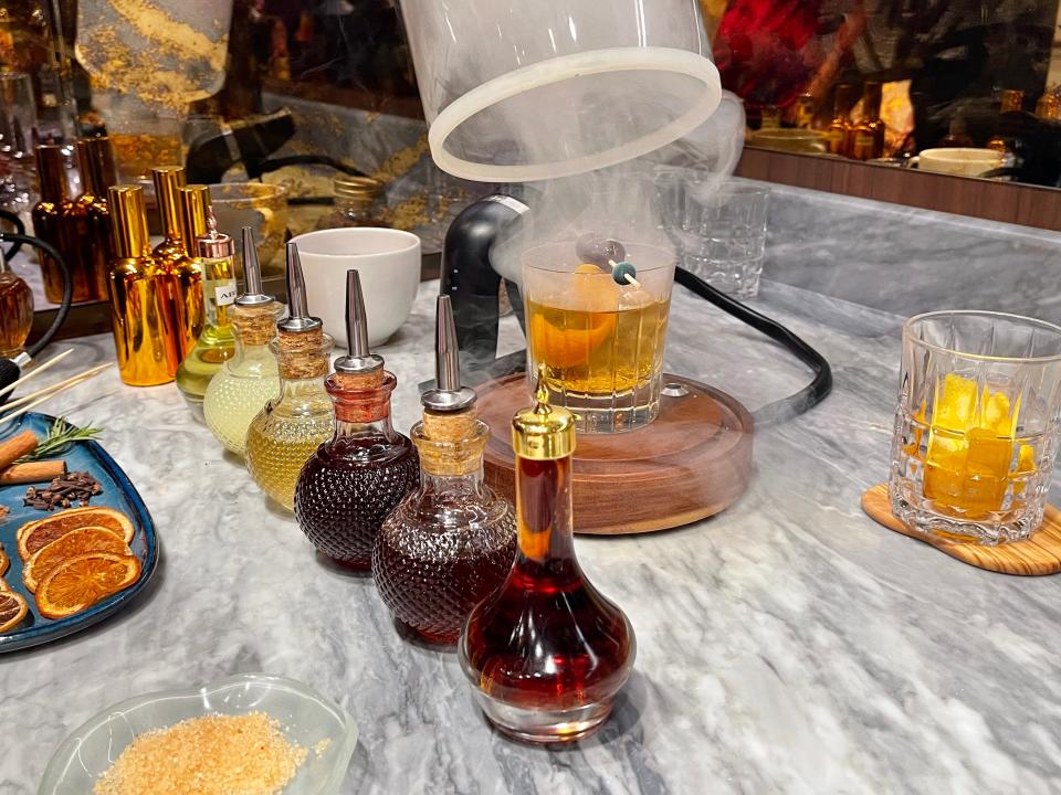 A cocktail being smoked on a bar with different colored liquid in bottles and garnishes on a plate.