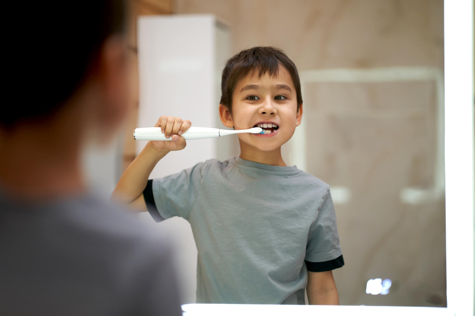 A boy brushes his teeth with an electric toothbrush near a backlit mirror.