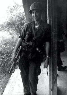 Daniel Ellsberg leans against a pole wearing army fatigues and holding a gun.