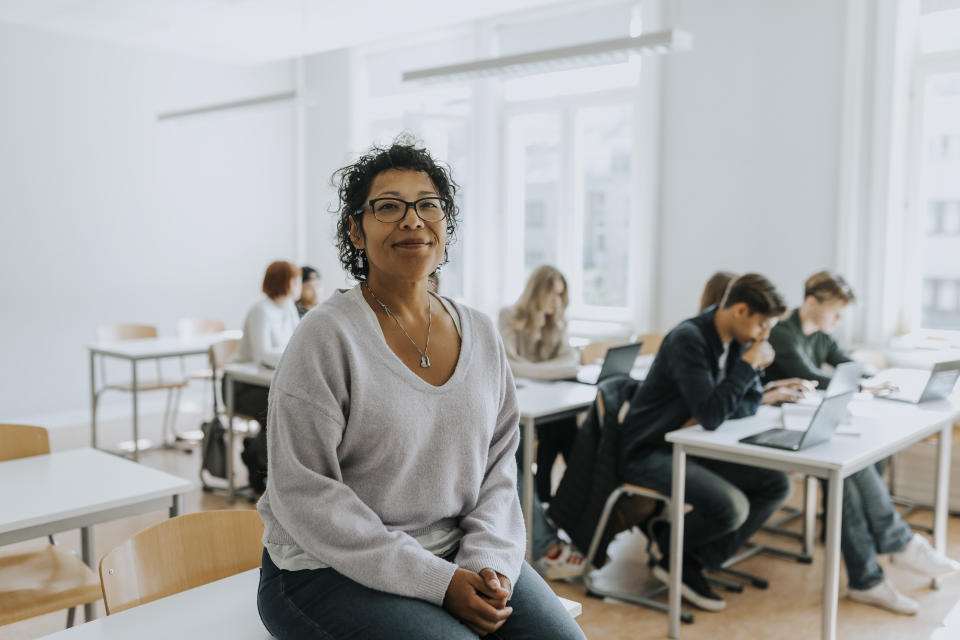 A person sits on a desk in a classroom with students working at tables in the background