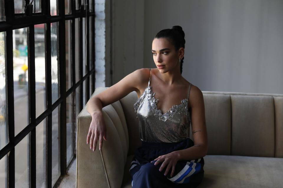 Dua Lipa sits on a couch by a window.