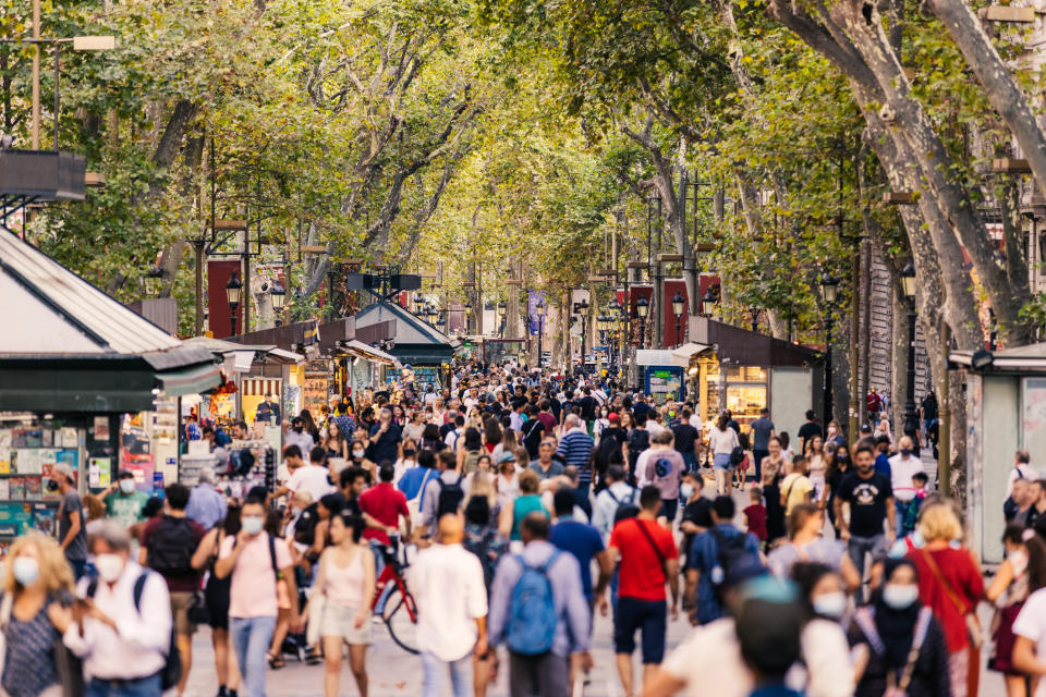 Crowded urban street with pedestrians walking, trees lining the path, and kiosks on the sides