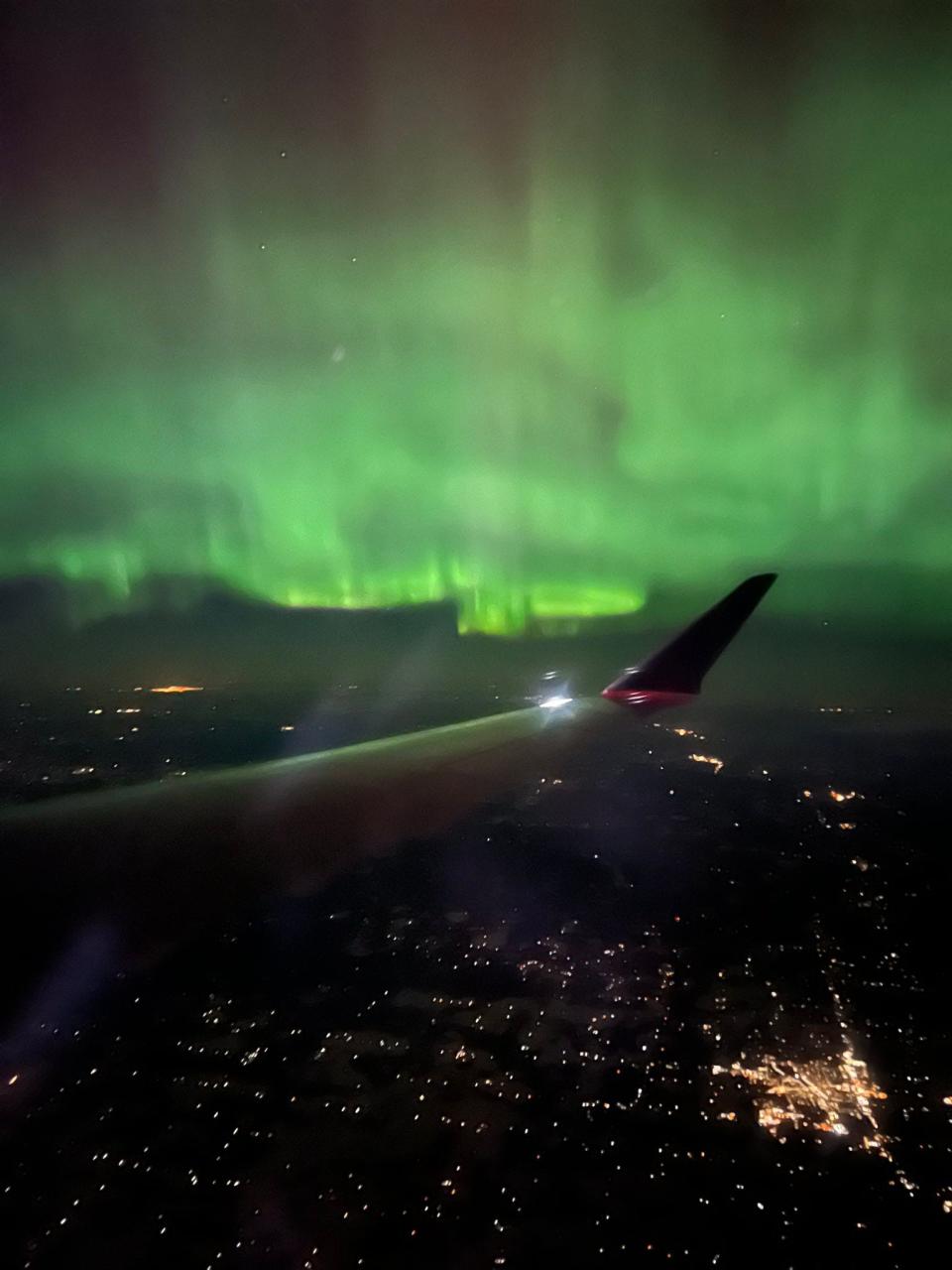 green aurora borealis northern lights cover the night sky above an airplane wing flying over a city