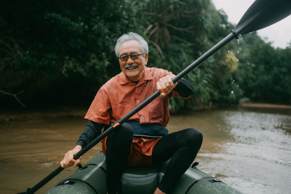 Elderly man, smiling, paddles a kayak on calm water surrounded by lush greenery. He wears a short-sleeve shirt over a long-sleeve top and athletic pants