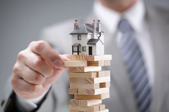 A businessman in a suit pulling out a wooden Jenga block, with a miniature home teetering at the top of the blocks.