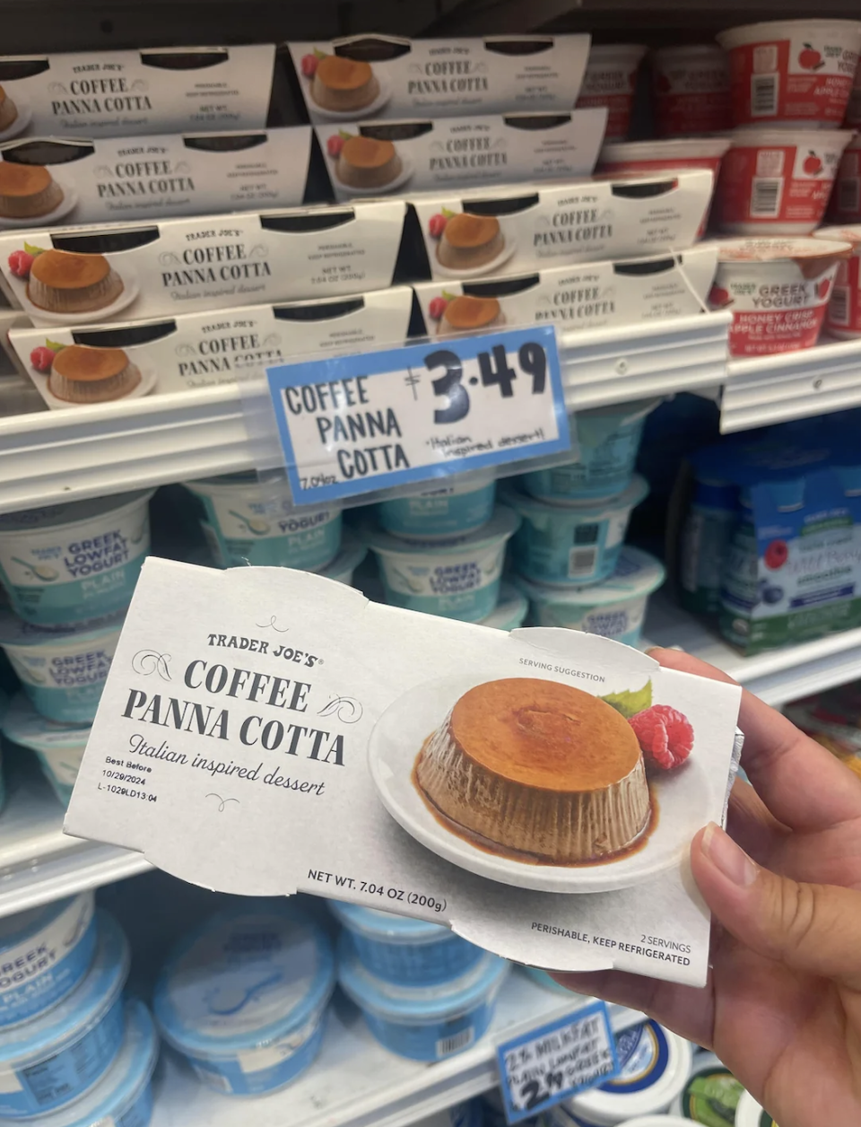 Hand holding a box of Trader Joe's Coffee Panna Cotta in front of a store shelf filled with more Coffee Panna Cotta and other Trader Joe's desserts. Price tag shows $3.49