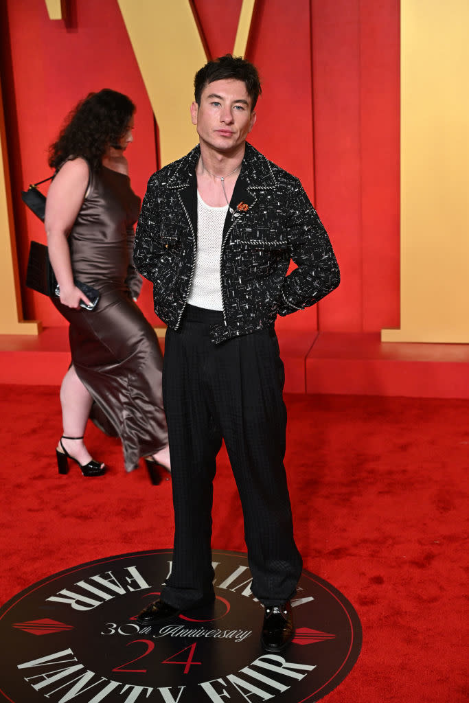 Man in a patterned jacket and trousers posing on event carpet; woman in background