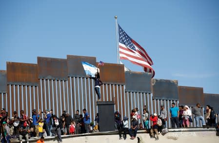 FILE PHOTO: A migrant, part of a caravan of thousands traveling from Central America en route to the United States, holds flags of Honduras and the United States in front of the border wall between the U.S. and Mexico in Tijuana