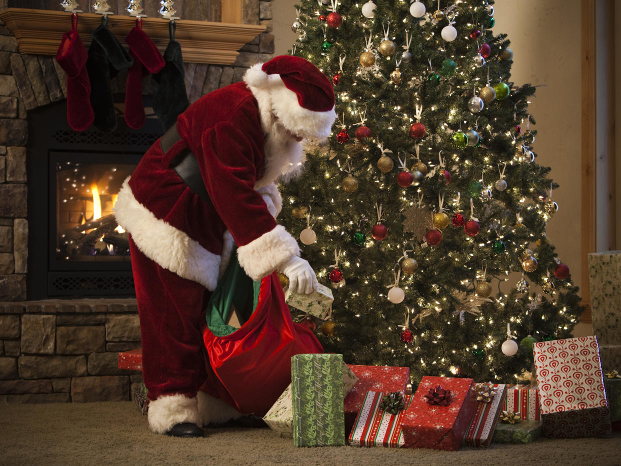 Santa Claus in a red suit putting wrapped presents beneath a decorated Christmas tree.