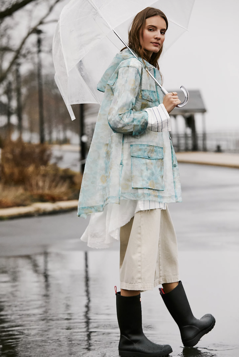 model in rain with umbrella wearing black rainboots, blue floral Festival Raincoat (photo via Free People)