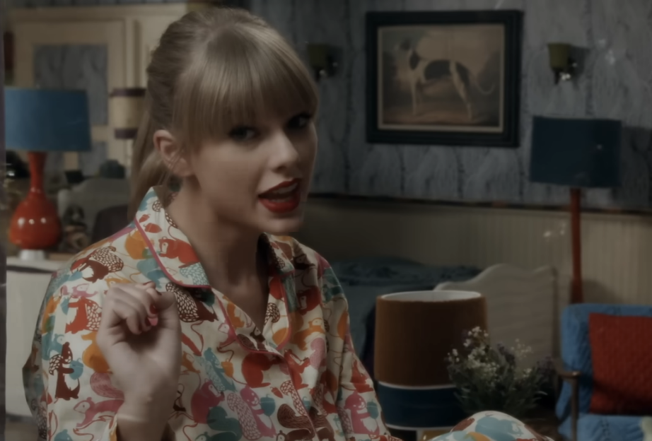 Taylor Swift in a patterned shirt, sitting indoors, looking surprised with mouth slightly open