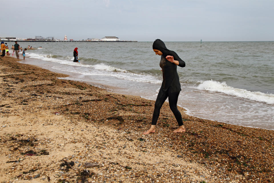 A young Muslim woman wearing a burkini  walks on Clacton beach, UK. Clacton pier in the the background.