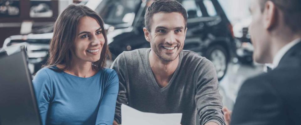 Young couple talking to car salesman at dealership, man is holding papers, everyone smiling