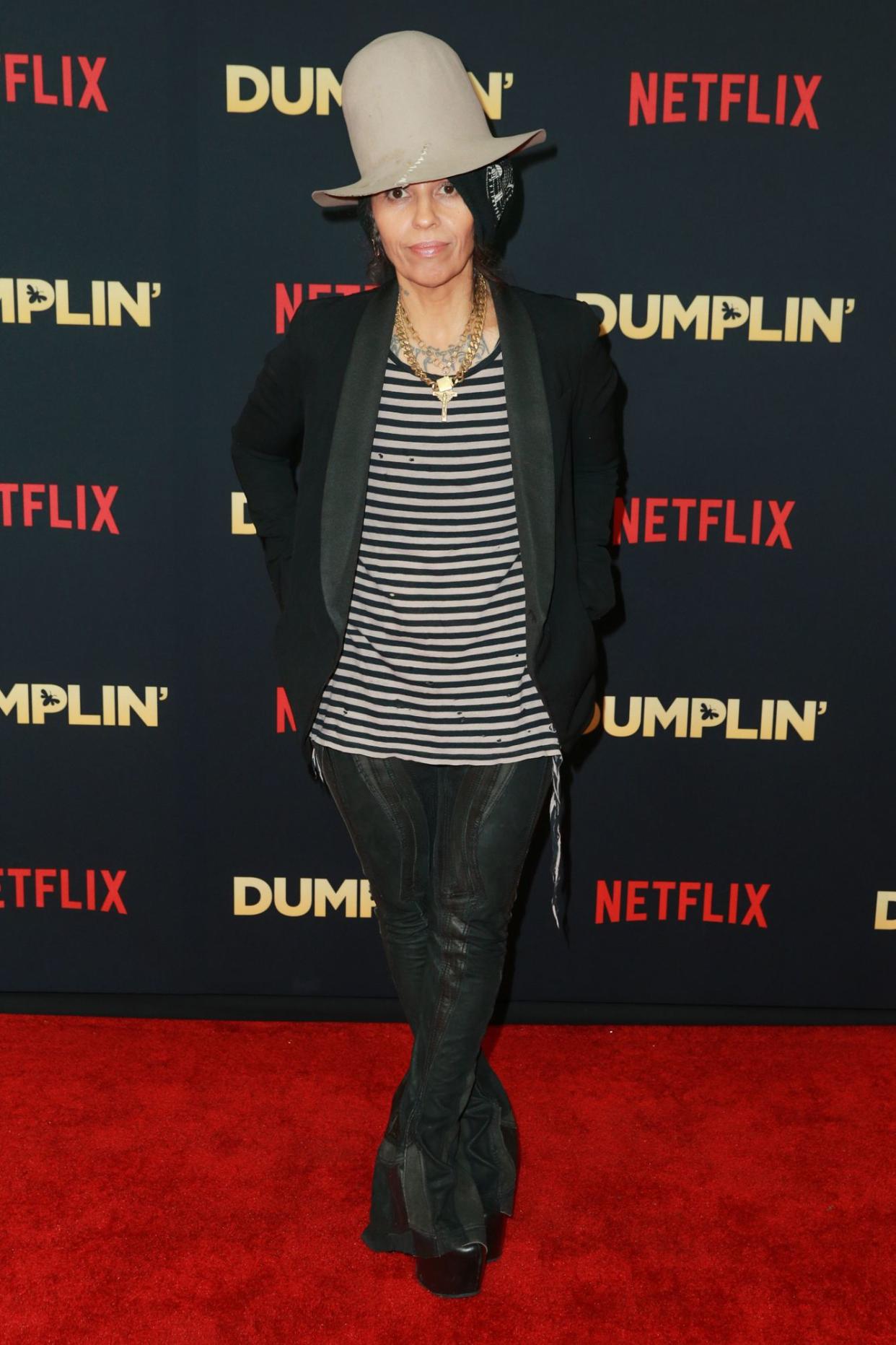 Linda Perry stands on a red carpet wearing a tall grey hat