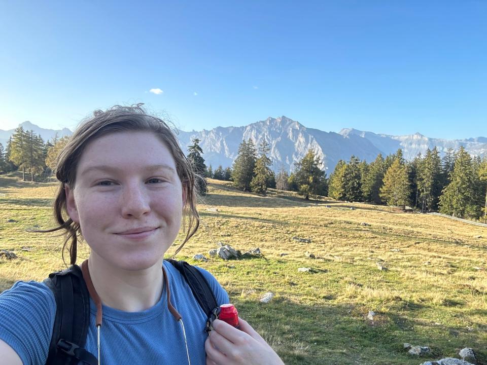 woman wearing backpack blue shirt taking selfie in front of mountain slope with peaks in background