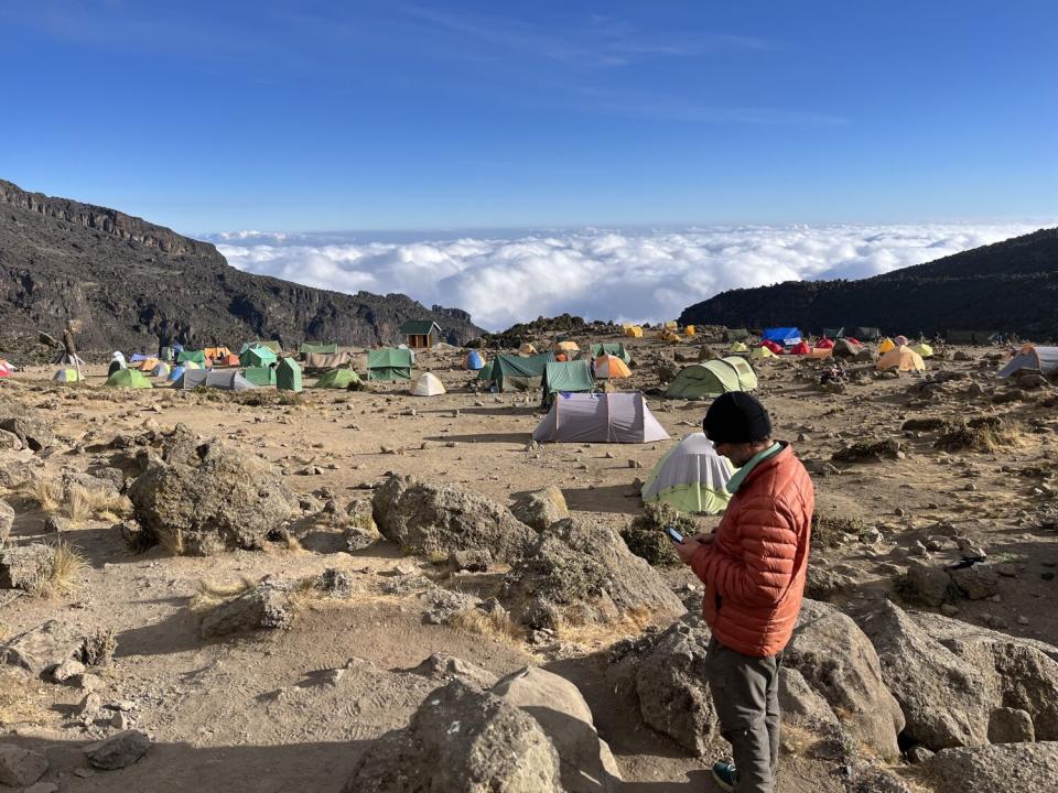 A man in an orange jacket and knit cap stands in a rocky mountainous landscape with tents in the background