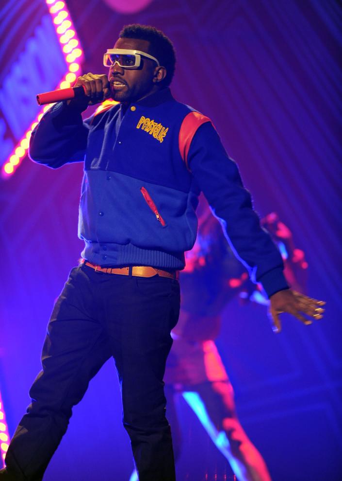 ye performing onstage at the 2008 amas, wearing a bright blue and red pastelle branded varsity jacket that's buttoned up. he's holding up a red microphone to his mouth, and wearing large white sunglasses