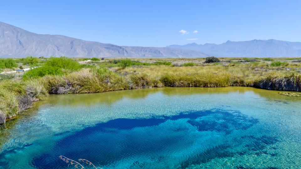 a blue green pool of water surrounded by grassland and shrubs with a blue sky above and high hills in the background.